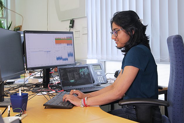 Frau an einem PC, auf einem Bildschirm Programmcode, auf dem anderen Messcharts.