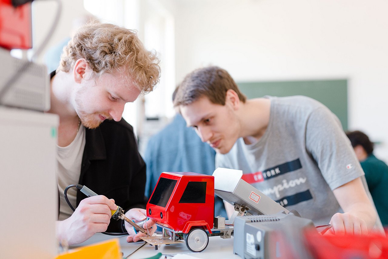 Am Stand der TU Ilmenau sind autonome Miniaturtransporter in Aktion. Erarbeitet wurden sie bei Orientierungsangeboten an der TU, die Praxis zu Beginn des Studiums vermitteln. Foto: TU Ilmenau/Stefan Riehmer. Zwei junge Männer an einem Tisch löten an einer Platine für einen LKW in Modellbaugröße