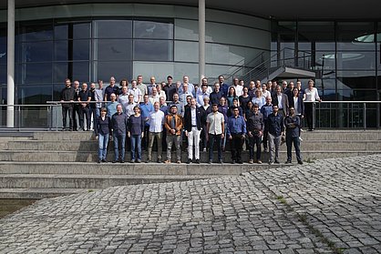 IMMS-Team in Ilmenau 2026. Insgesamt arbeiten rund 100 Personen am IMMS in Ilmenau und Erfurt. Foto: IMMS. Rund 60 Männer und Frauen stehen in mehreren Reihen auf einer Treppe vor einem großen Gebäude mit Glasfassade.