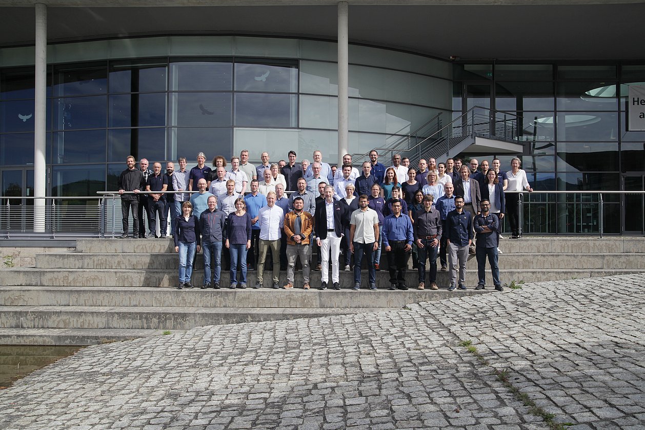 IMMS-Team in Ilmenau 2026. Insgesamt arbeiten rund 100 Personen am IMMS in Ilmenau und Erfurt. Foto: IMMS. Rund 60 Männer und Frauen stehen in mehreren Reihen auf einer Treppe vor einem großen Gebäude mit Glasfassade.