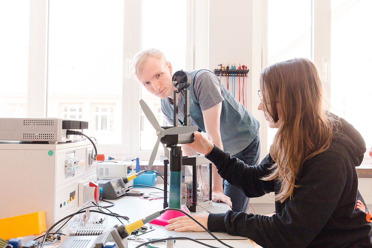 In den Orientierungsangeboten an der TU Ilmenau kann man schon zum Studienstart in die Praxis eintauchen, wie zum Beispiel beim Bau eines Windkraftanlagen-Modells, das auch zur Langen Nacht getestet werden kann. Foto: TU Ilmenau/Stefan Riehmer. Eine junge Frau und ein Junger Mann arbeiten an einem Windkraftanlagen-Modell mit Rotor, Generator und Platinen, daneben Lötkolben und Messgeräte.