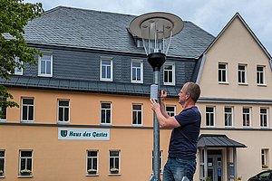 Mann auf einer Leiter schraubt einen Sensor an einen Lampenmast vor einem Haus.