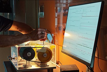 Edge-AI-based monitoring with “AWARE” (Advanced wireless AI-enabled real-time environment). This allows, for example, a gearbox to be monitored for wear and defect. Photograph: IMMS. A gearbox is adjusted using an inner hex key. A monitor displays the readings.