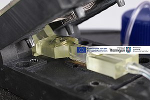 Untersuchungsaufbau mit Klappfassung, darin der GraFET-Chip, darüber eine mikrofluidische Flusszelle und darüber die Kontaktiernadeln für den Chip.