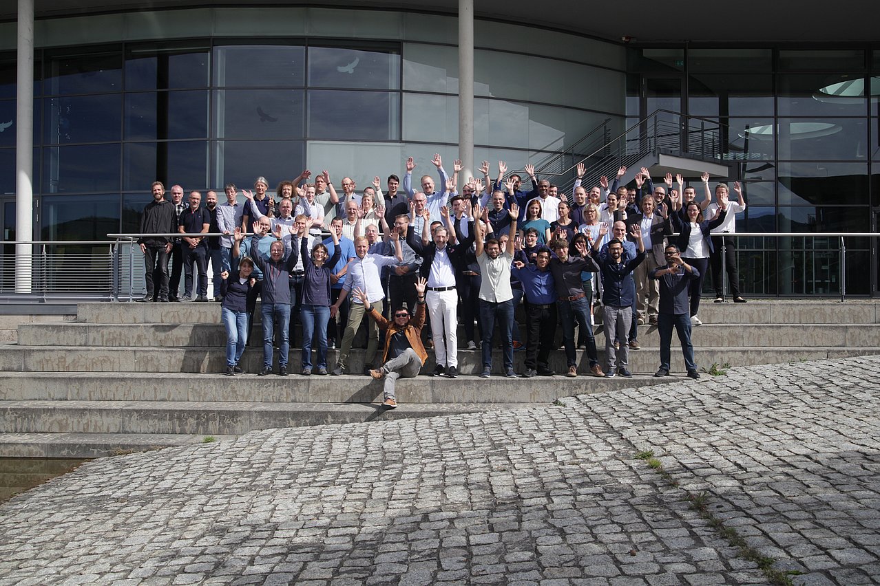 IMMS team in Ilmenau 2026. A total of around 100 people work at IMMS in Ilmenau and Erfurt. Photograph: IMMS. Around 60 men and women are standing in several rows on a flight of steps in front of a large building with a glass facade.