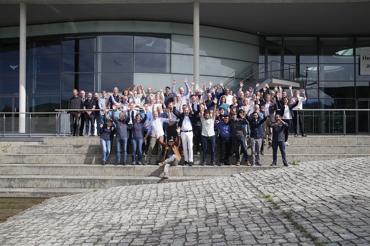 IMMS-Team in Ilmenau 2026. Insgesamt arbeiten rund 100 Personen am IMMS in Ilmenau und Erfurt. Foto: IMMS. Rund 60 Männer und Frauen stehen in mehreren Reihen auf einer Treppe vor einem großen Gebäude mit Glasfassade.