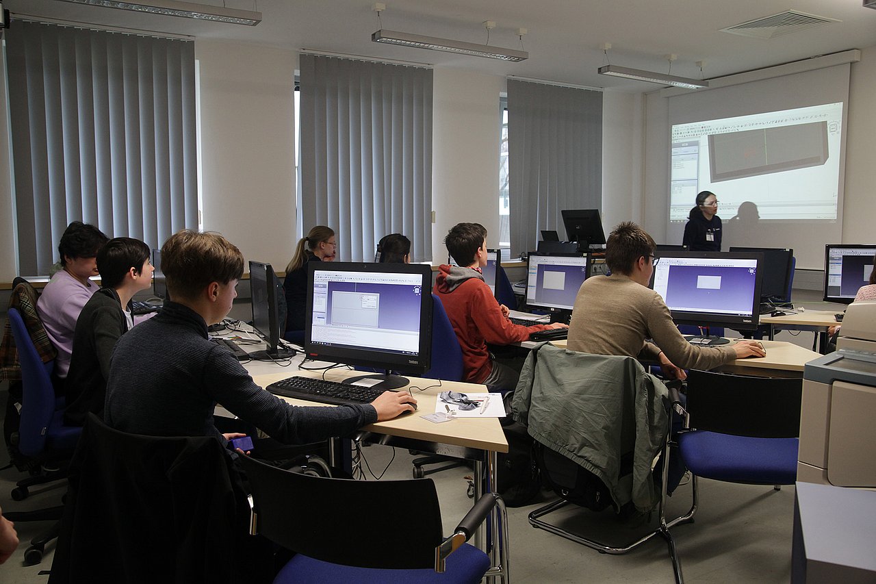 Einblick in die Konstruktion für z.B. 3D-Druck zum Forschungstag am IMMS. Foto: IMMS. Eine Gruppe von Schülerinnen und Schülern vor PC-Monitoren wird von einer Mitarbeiterin des IMMS beim Bedienen des Konstruktionsprogramms unterstützt.