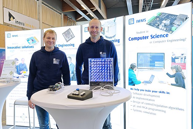 Among other things, IMMS presents its opportunities for work placements and thesis projects at careers fairs and provides an insight into what it is like to work at the institute. Photograph: IMMS. Two men in front of an exhibition stand, behind a bar table with electronic equipment.