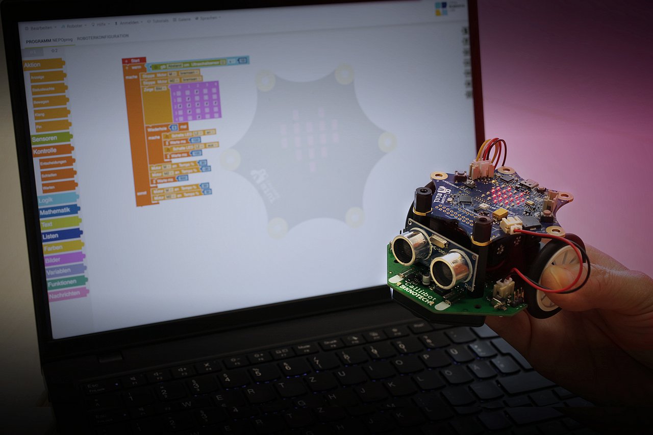 Solche Calli:bots können zur Langen Nacht programmiert und zum Fahren auf die Teststrecke geschickt werden. Foto: IMMS. Ein handtellergroßer Fahrroboter mit freiliegenden Platinen, LEDs und Kabeln in einer Hand vor einem Laptop, der die für Kinder geeignete grafische Programmieroberfläche zeigt.