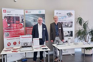 Zwei Personen vor drei RollUps und hinter zwei Tischen mit Infomaterial und verschiedenen Demonstratoren. 