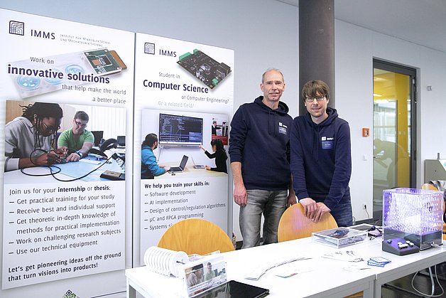 Two people next to an exhibition wall and behind a table with electronics and information material. 